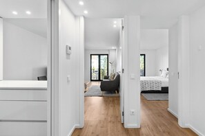 Een modern interieur met een gang die leidt naar een slaapkamer en een woonruimte, met een houten vloer, witte muren en een groot raam.