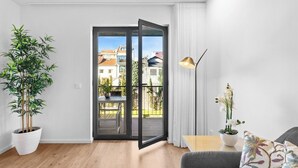 Una sala moderna con un amplio ventanal corredero que da a un balcón, una planta en maceta, una mesa con una lámpara y un sofá con cojines decorativos.