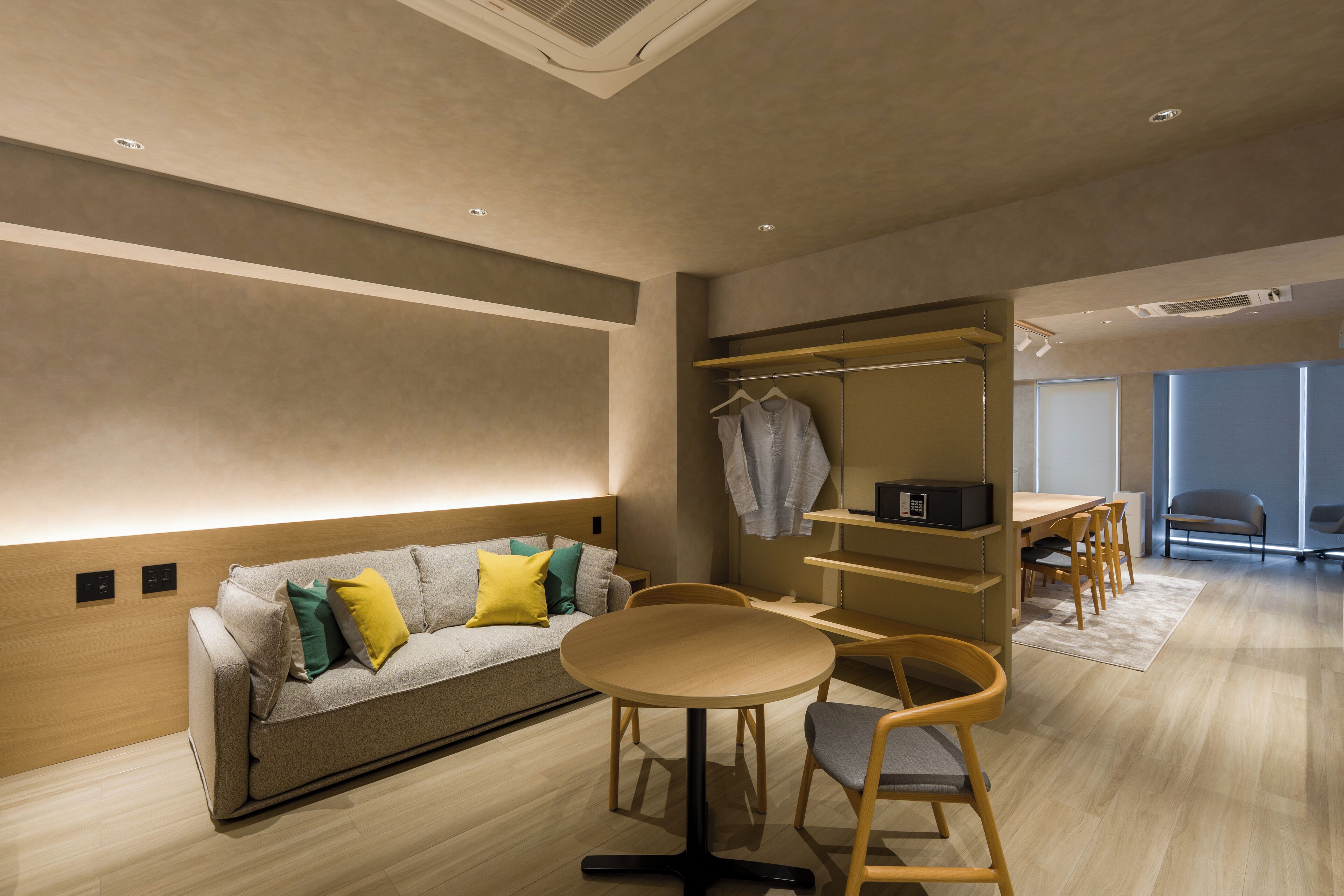 Une chambre d’hôtel moderne avec un canapé, une table ronde et des chaises. On y trouve un porte-manteau avec des vêtements suspendus et une petite étagère sur laquelle est posé un micro-ondes.