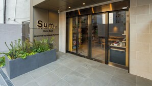 A modern building with a glass entrance, a sign reading "Sumu Ueno East powered by Airbnb Partners," and a planter with greenery in front. - Sumu Ueno East (Tokyo)