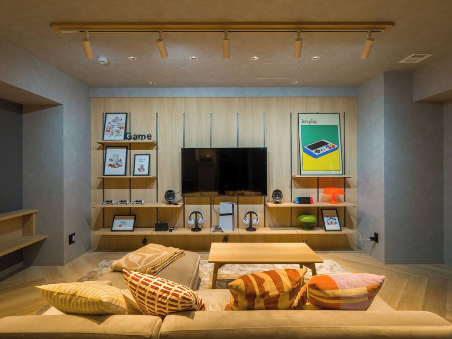 A modern living room with a large flat-screen TV, wooden shelves with framed pictures, and a comfortable seating area with cushions.