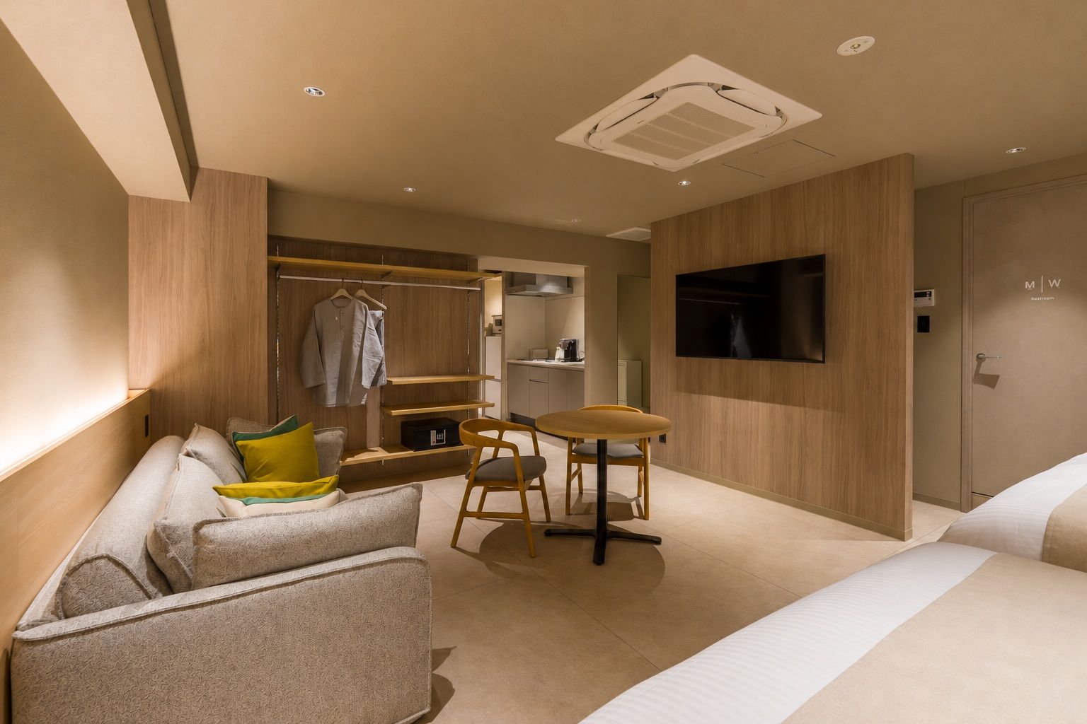 A modern hotel room with a bed, a seating area with a table and chairs, a TV mounted on the wall, and a wardrobe.