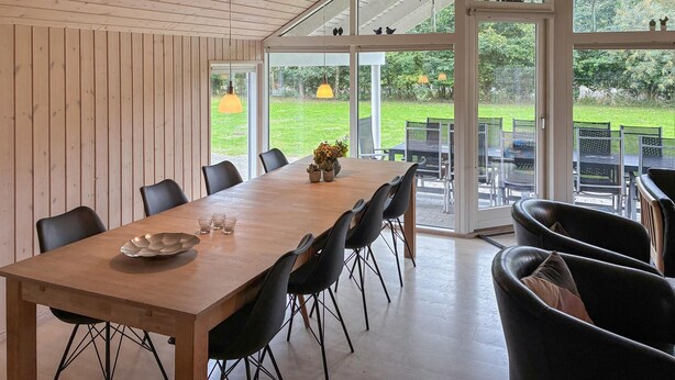 A dining area with a long wooden table, black chairs, and a glass door leading to an outdoor patio with chairs and tables. - Beautiful home in Idestrup with sauna (Idestrup)