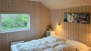 A bedroom with wooden walls, a bed with white bedding, a window with a view of greenery, a bedside lamp, and a wall art piece. - Beautiful home in Idestrup with sauna (Idestrup)