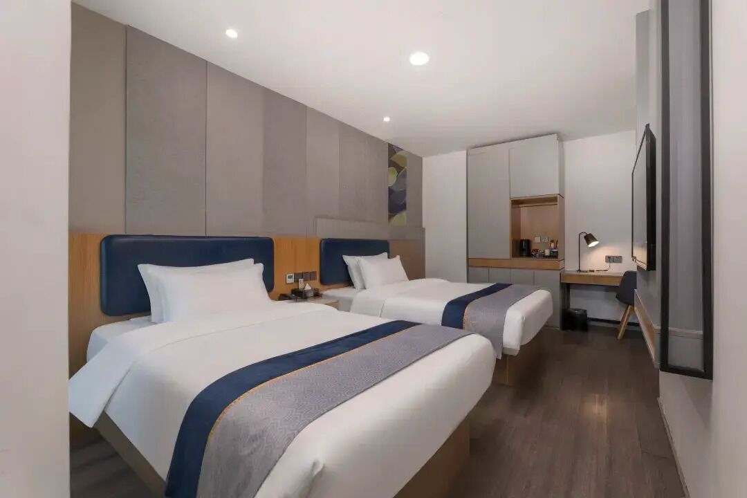 A hotel room with two beds, a desk, a mirror, and a lamp.