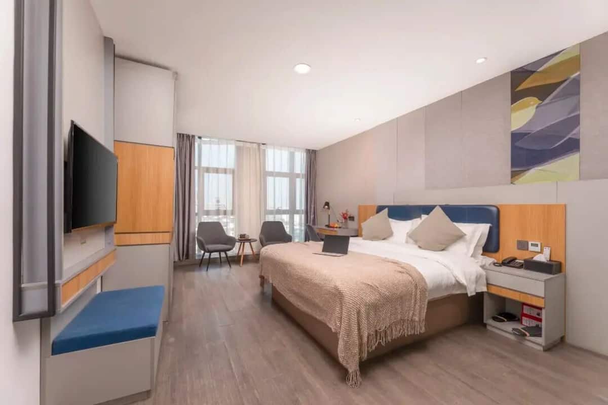 Una camera d'albergo moderna con un letto grande, un comodino e una TV a schermo piatto. Si vede anche una scrivania con una sedia e una finestra con tende.