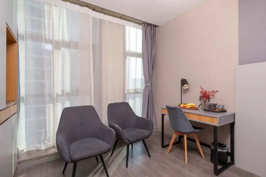 Une petite pièce moderne, avec deux fauteuils gris, un bureau doté d’une lampe et une fenêtre ornée de rideaux légers.