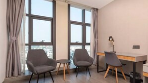A modern room with two grey armchairs, a small round table, a desk, and a lamp. Large windows with curtains are on the left, and a city view is visible. - suzhouzhexujiajiudian (Suzhou)