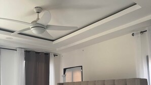 Ein modernes Schlafzimmer mit einer gepolsterten Kopfstütze, einem großen Bett mit gemusterten Betttextilien, einem Deckenventilator und einem Fenster mit Vorhängen.
