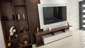 Ein modernes Wohnzimmer mit einem großen Flachbildfernseher an einer weißen Wand, Holzregalen mit Dekoration und einer eleganten Unterhaltungseinheit.
