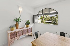 Ein Essbereich mit einem Holztisch und Stühlen, einer Schrankwand mit Ziegeloptik und Blumentöpfen sowie einem Fenster mit rundem Bogen, das auf einen Garten hinausblickt.