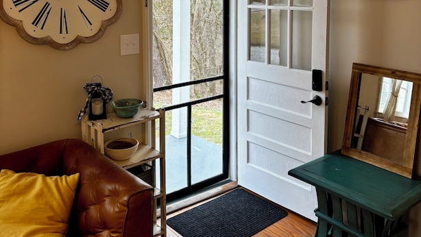 A room with a wooden door, a clock, a brown leather couch, a small table, and a view through the door. - Quiet, Private Home in Kingston Springs for Two (Kingston Springs)