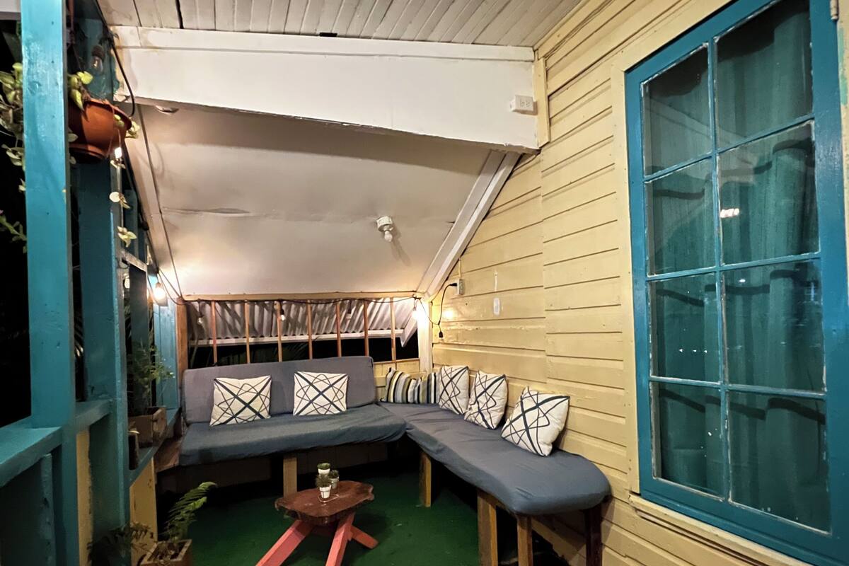 Un porche confortable et fermé, doté d’un plafond en bois, d’un banc bleu avec des coussins à motifs et d’une petite table en bois.