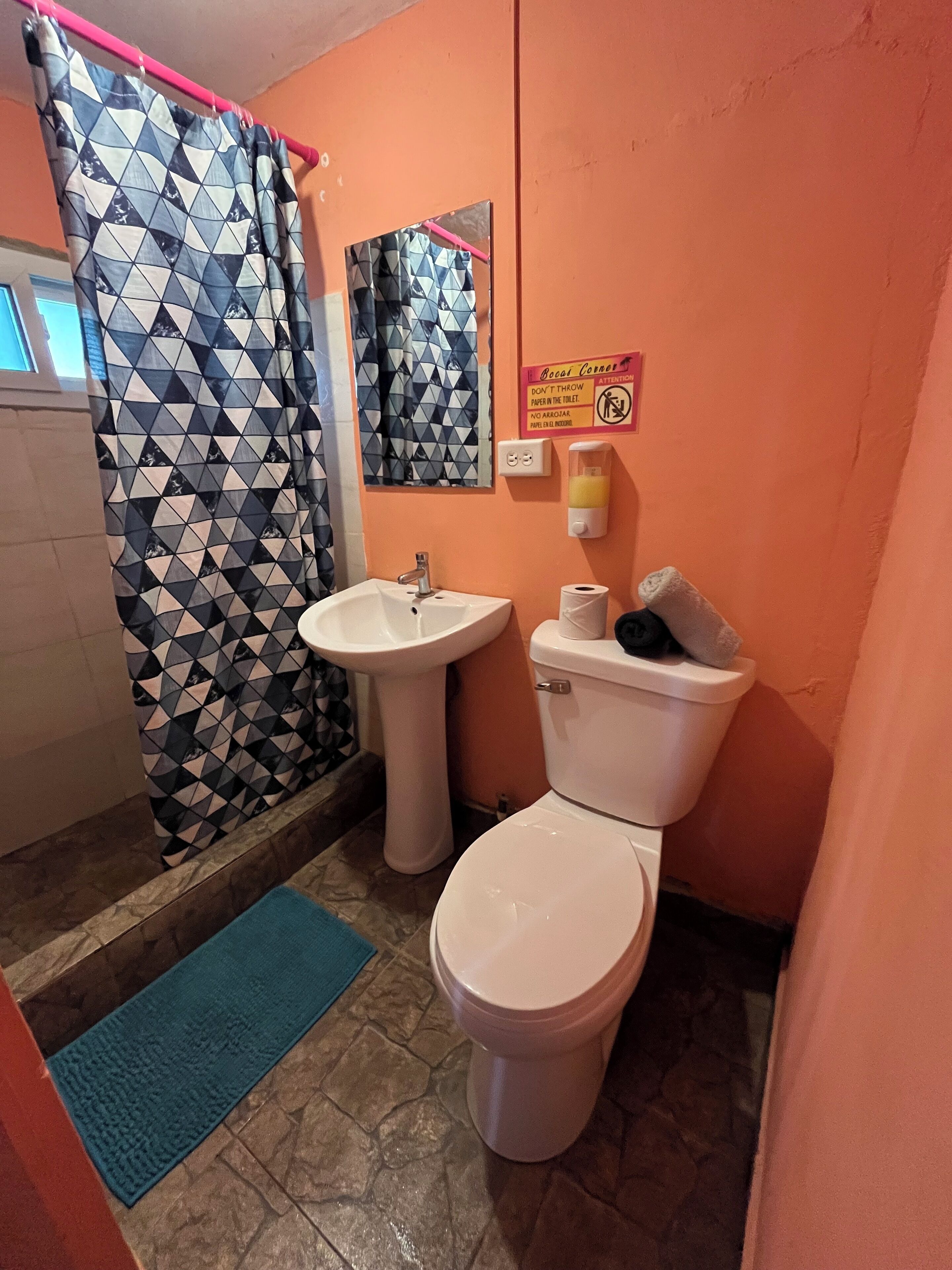 A bathroom with a shower curtain, sink, toilet, and a hand sanitizer dispenser.