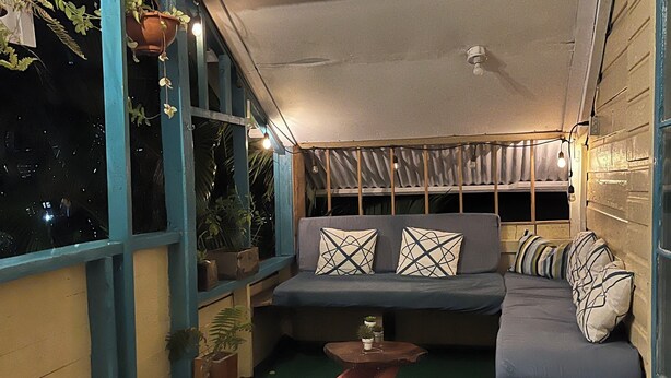 A cozy, enclosed porch area with a wooden bench and a cushioned seating area, featuring a small table and potted plants. - The Bocas Corner Hostel (Bocas del Toro)