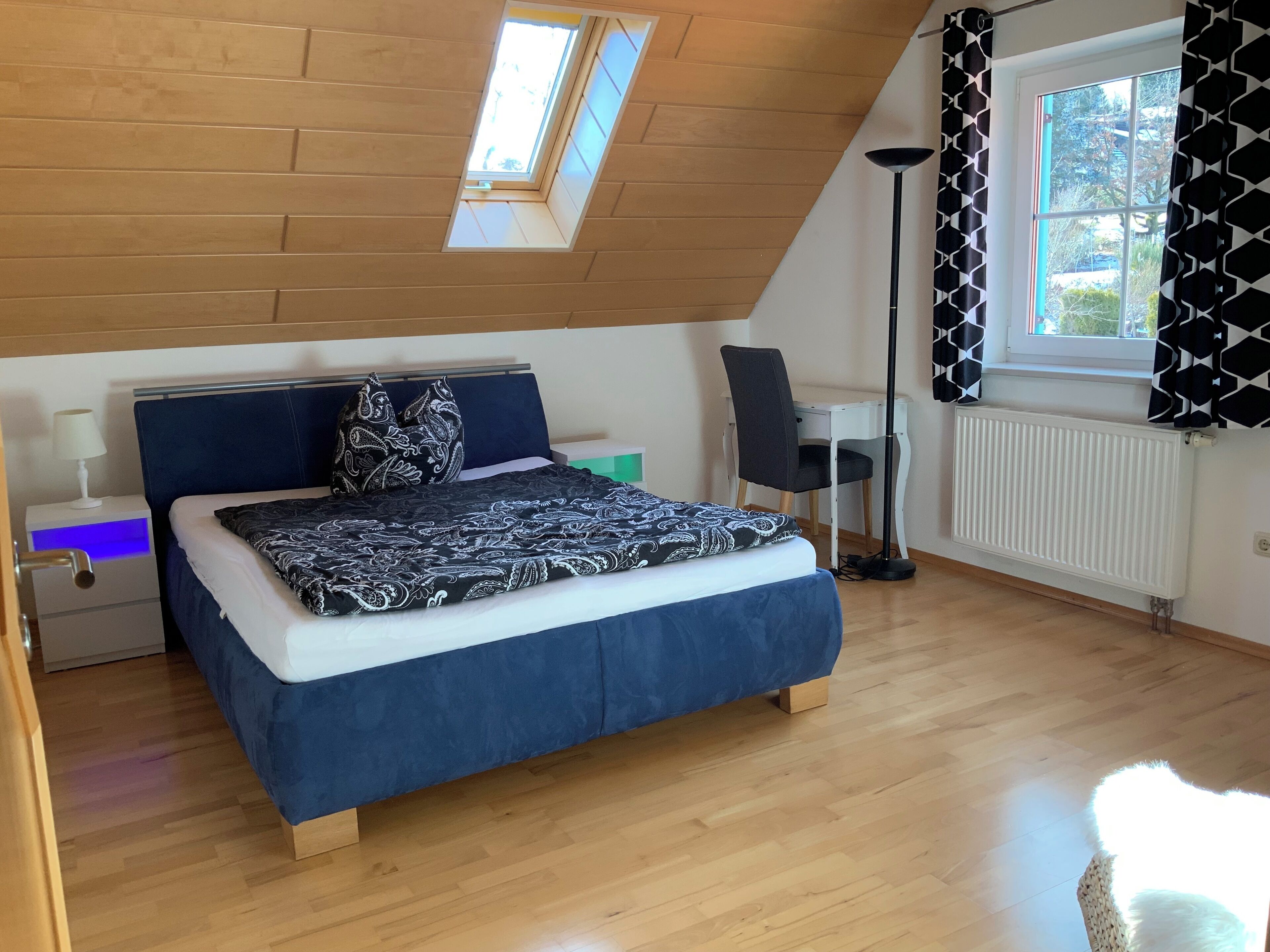 A bedroom with a sloped ceiling, a bed with a patterned bedspread, a desk with a chair, a standing lamp, and a window with patterned curtains.