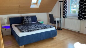 Une chambre avec un plafond en pente, un lit recouvert d’une couvre-lit à motifs, un bureau avec une chaise, un lampadaire et une fenêtre ornée de rideaux à motifs.