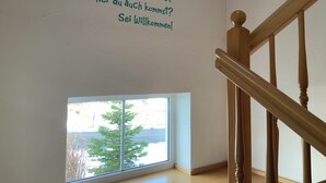 Une pièce avec un escalier en bois, une petite fenêtre donnant sur les arbres et un mur orné d’un message de bienvenue en allemand.