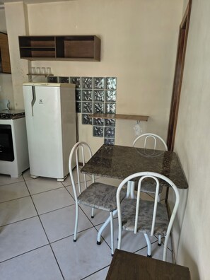 A small kitchen with a white refrigerator, a stove, and a tiled backsplash. There is a table with four chairs and a wooden stool. - Comfortable ground-floor apartment facing the street, 10 minutes from the beautiful Itaparica beach. (Vila Velha)