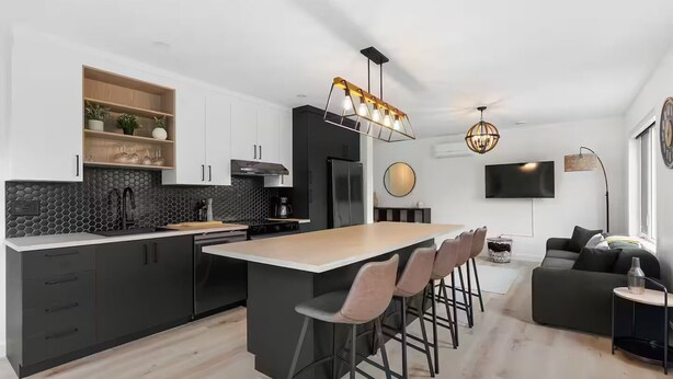 A modern kitchen with a central island, bar stools, a dining table, and a living area with a sofa and TV.