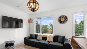 A modern living room with a black sofa, a flat-screen TV, a large window, and a clock on the wall.