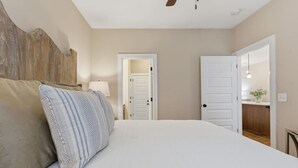 Une chambre à coucher avec un lit, des oreillers, une table de chevet, une lampe et une porte donnant sur une autre pièce.