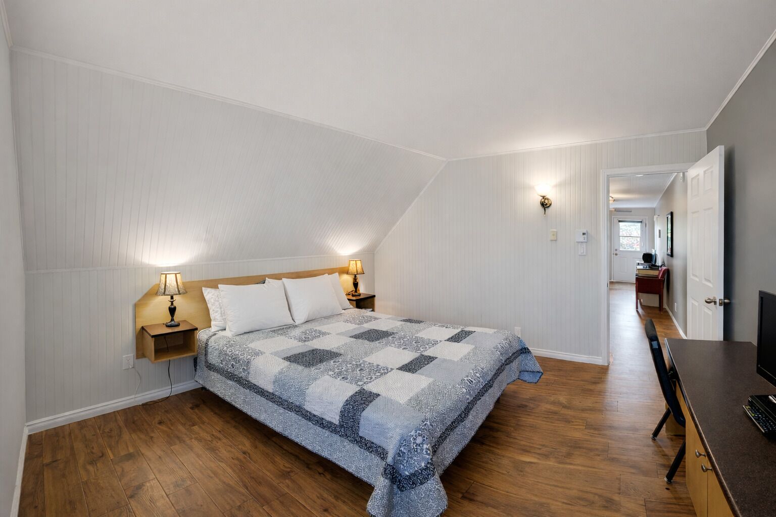 A bedroom with a wooden bed, two bedside tables with lamps, a desk with a chair, and a TV.