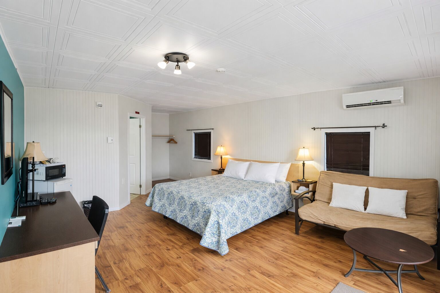 A hotel room with a bed, a sofa, a desk with a lamp, a small table, and an air conditioning unit.
