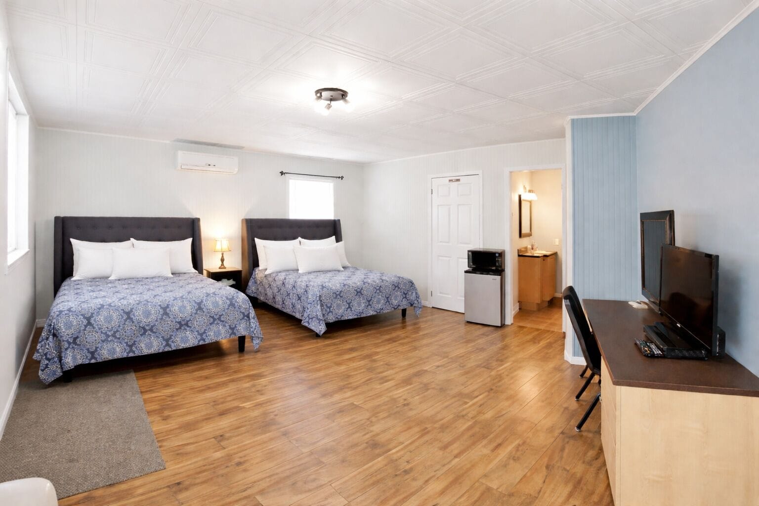 Habitación de hotel con dos camas, un escritorio con televisor, una pequeña cocina y la puerta del baño.