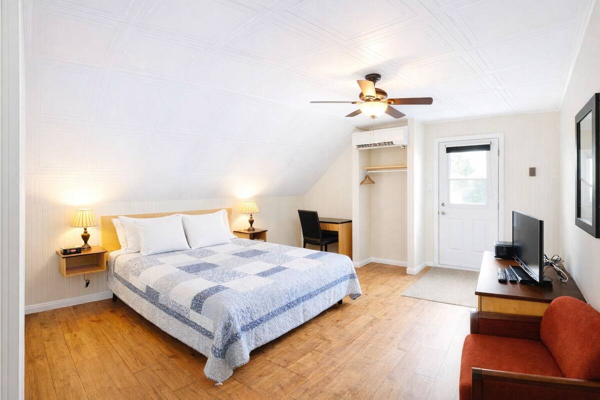 A bedroom with a bed, bedside lamps, a ceiling fan, a desk with a computer, a chair, and a wardrobe.