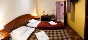A hotel room with a wooden headboard, a bed with a plaid bedspread, a nightstand with a lamp, a small table with a vase, and a chair. - Hotel Tamaris Chiclayo (Chiclayo)