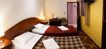 Una habitación de hotel con cabecera de madera, cama con colcha escocesa, mesita de noche con lámpara, una pequeña mesa con un florero y una silla.