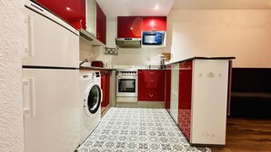 Une cuisine de taille réduite, équipée d’un lave-linge, d’un four et de meubles de rangement rouges.