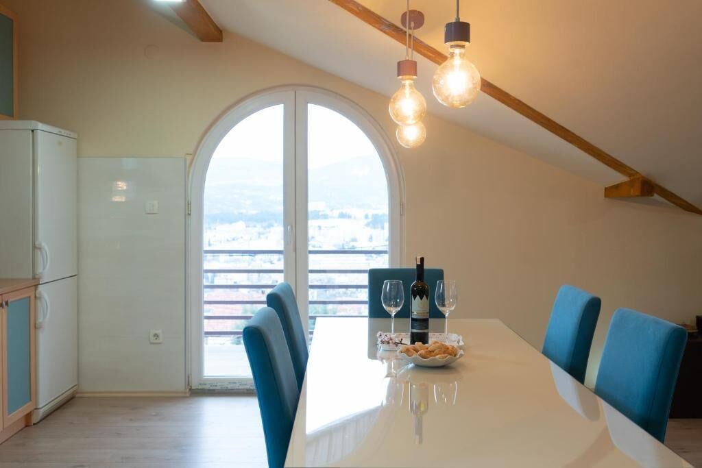A dining area with a white table, blue chairs, a bottle of wine, and glasses. There is a view through a large arched window.