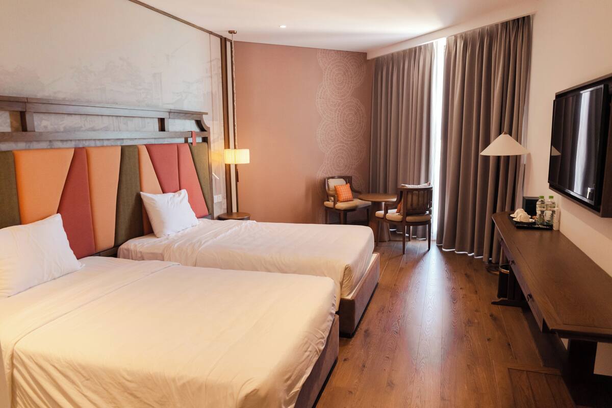 Et hotelværelse med to senge, et skrivebord, en stol, et fjernsyn og et vindue med gardiner.