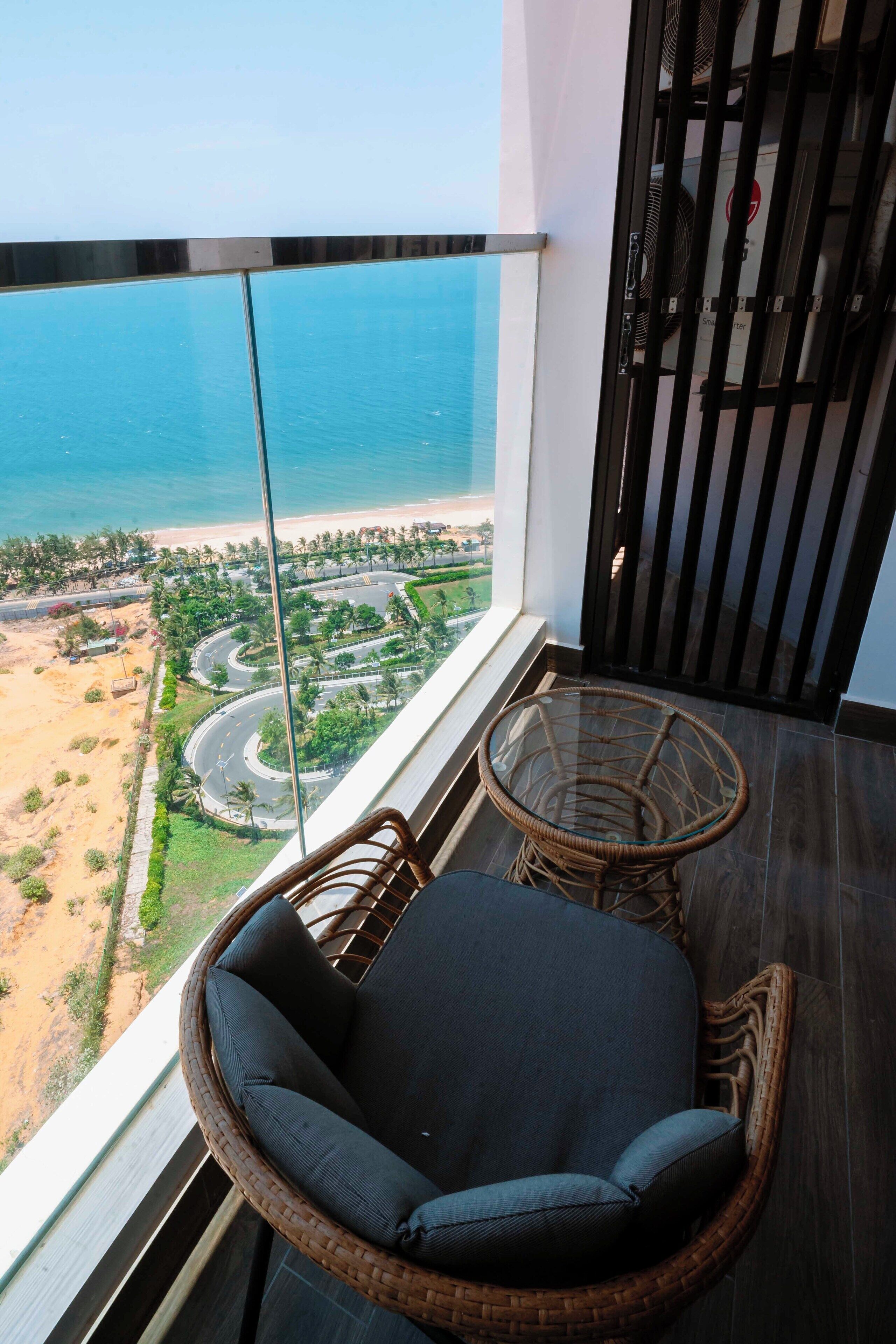 A balcony with a wicker chair and a small table, overlooking a beach and winding roads.
