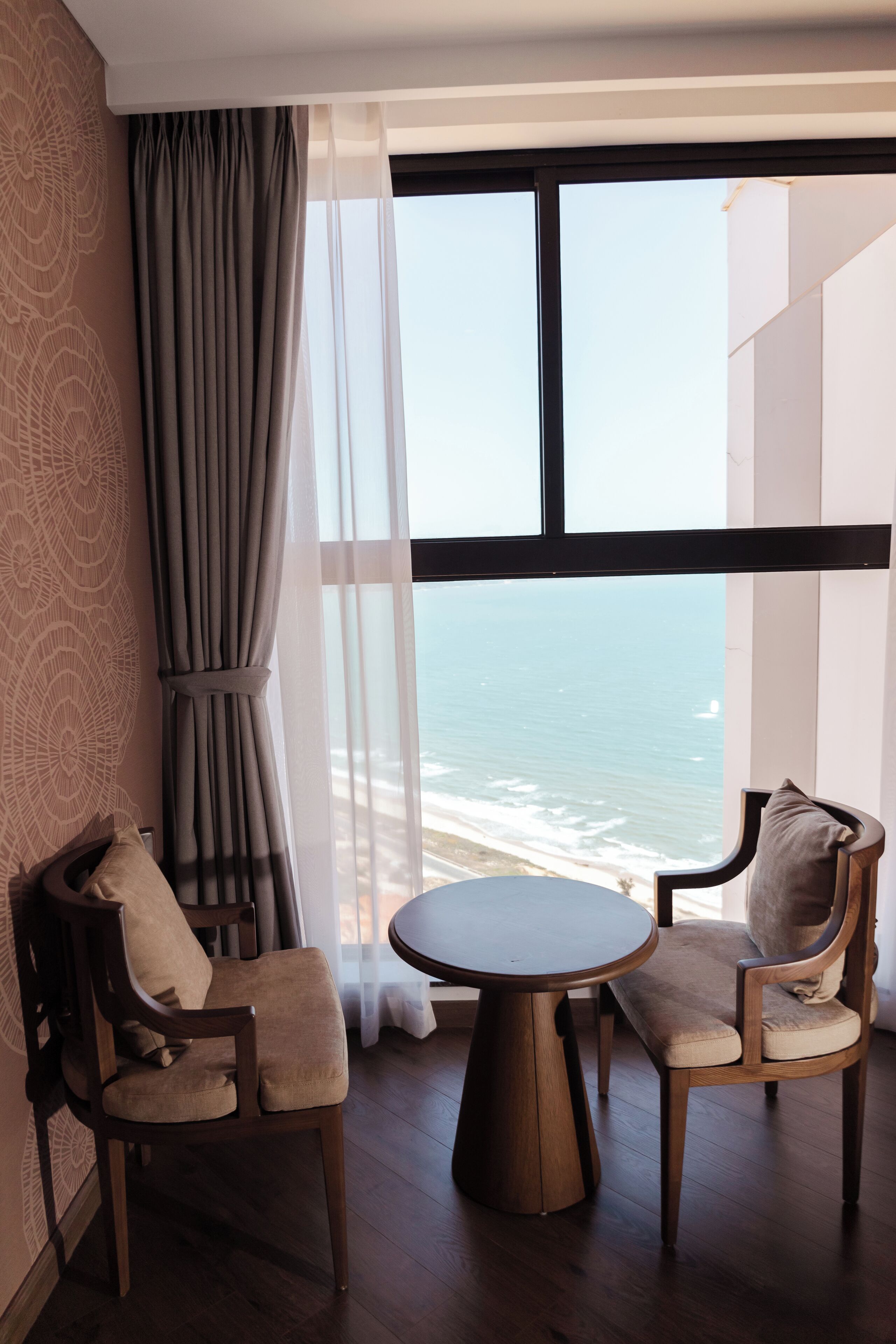 A room with a round wooden table and two chairs, a large window with a view of the ocean, and a curtain on the window.