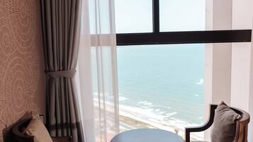 A room with a round wooden table and two chairs, a large window with a view of the ocean, and a curtain on the window.