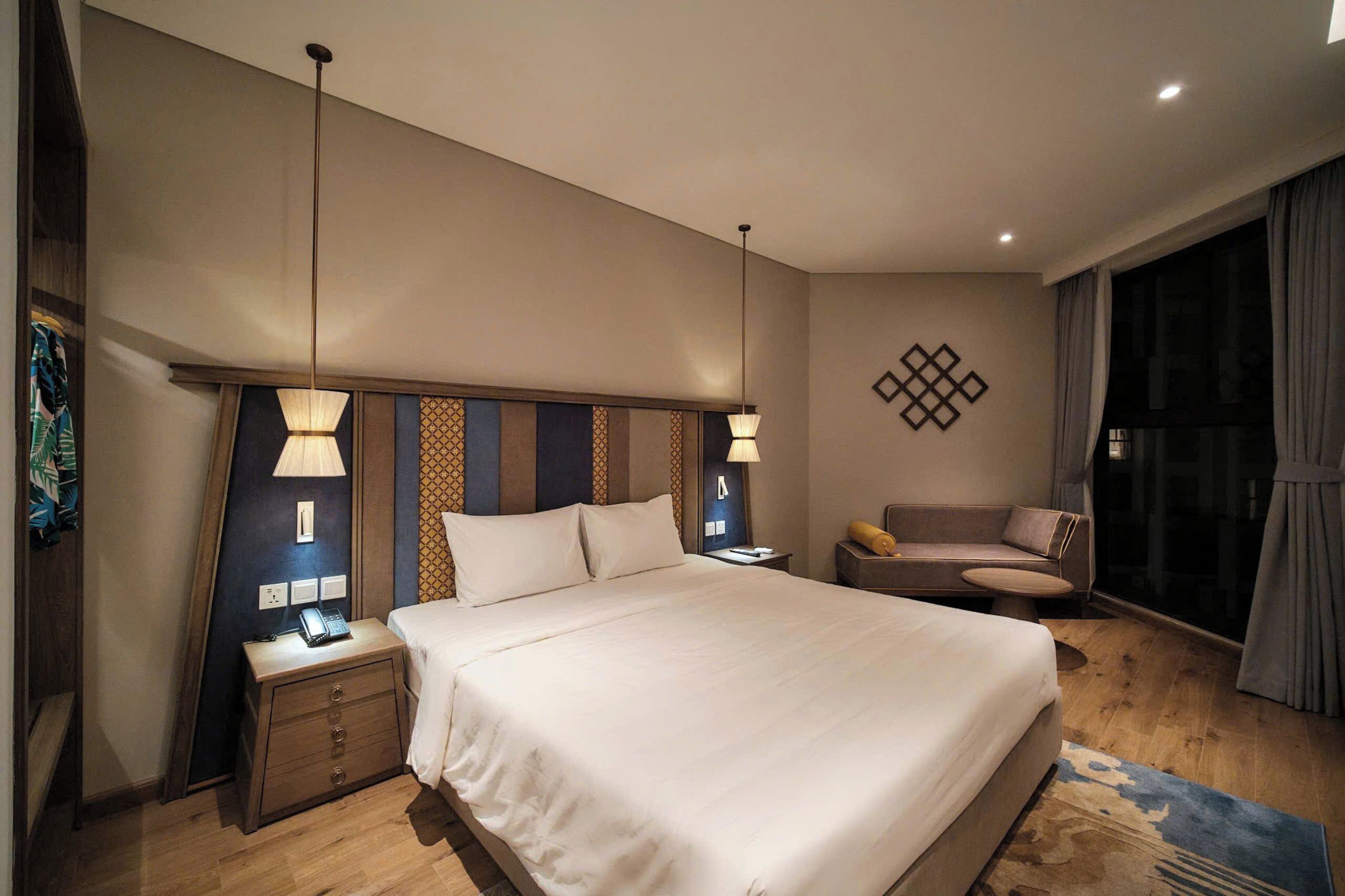 Habitación de hotel con una cama grande, lámparas de noche, un pequeño sofá, una mesita de noche y un adorno en la pared.