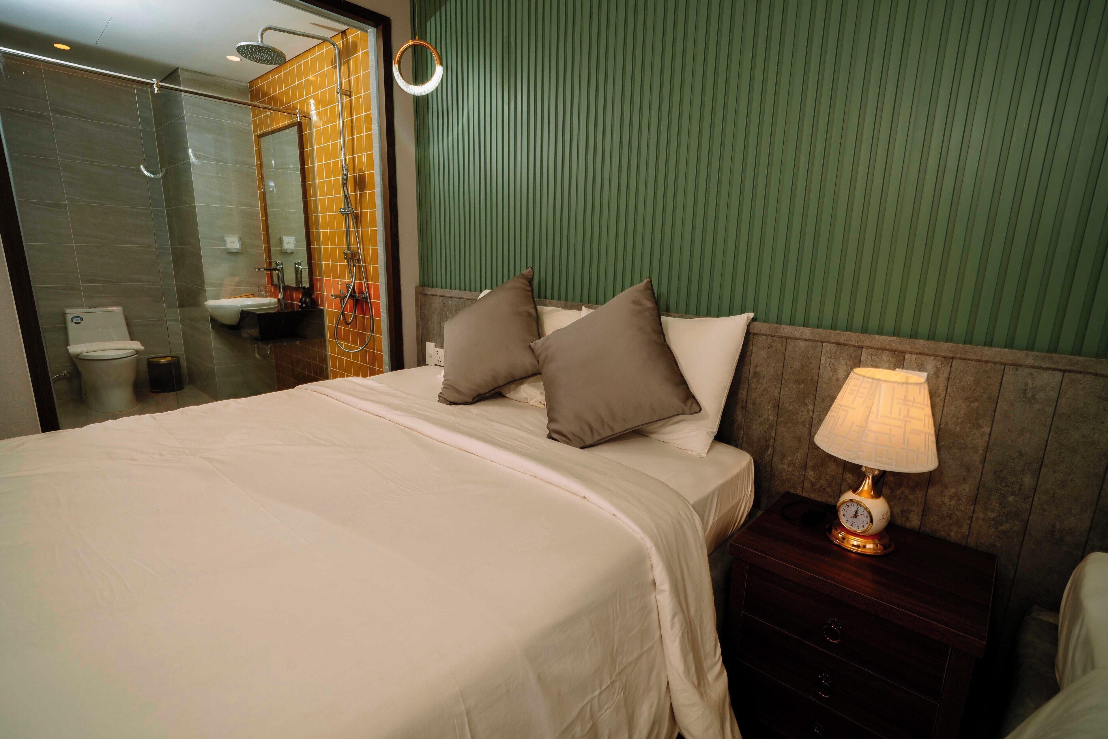 A hotel room with a bed, a nightstand with a lamp, a mirror, and a bathroom with a shower and toilet.