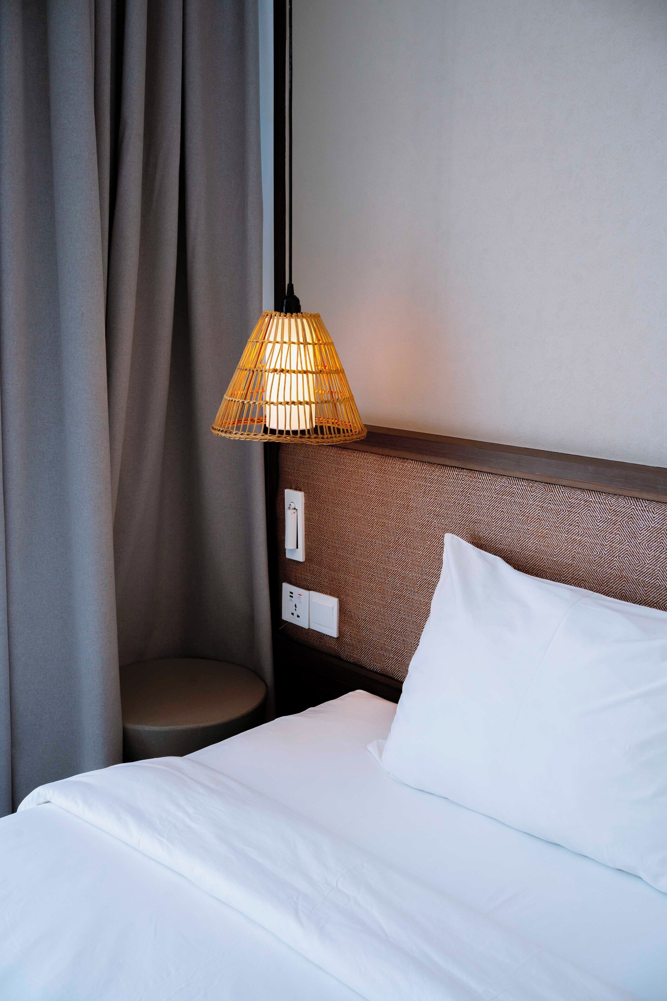 Een net opgemaakt bed met witte lakens, een gestructureerd hoofdbord en een geweven lamp.