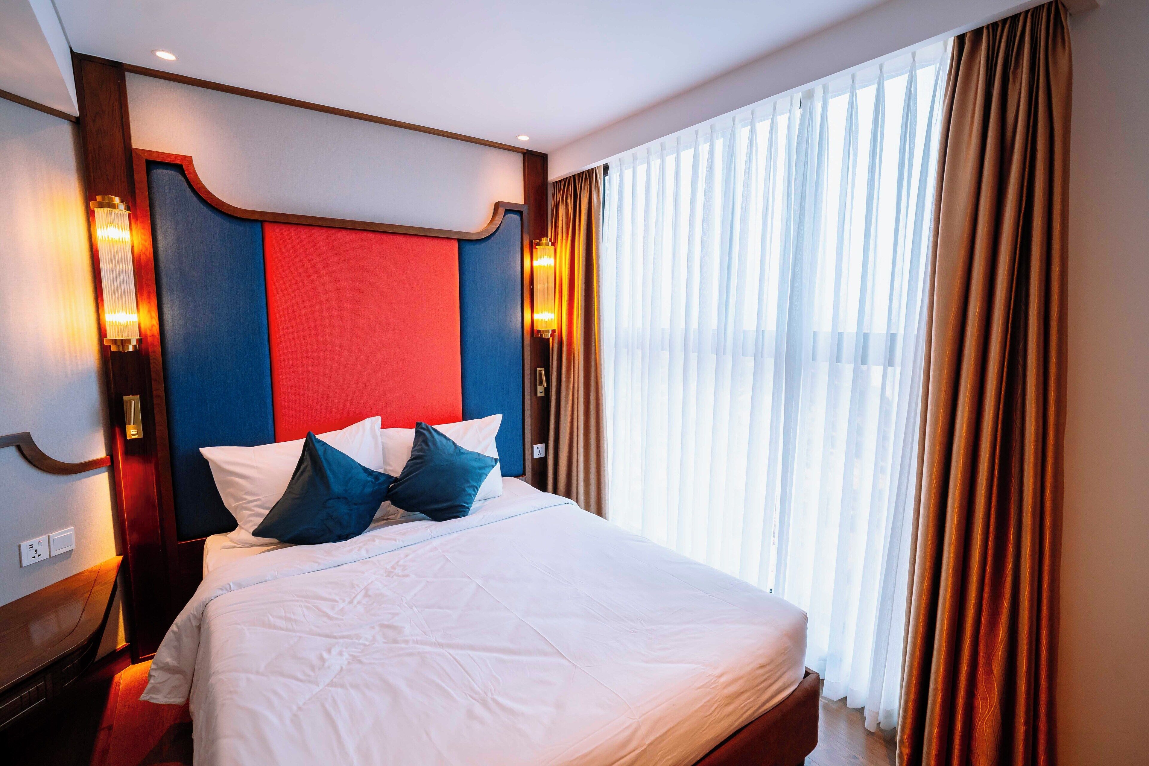 A hotel room with a large bed, a red and blue headboard, and a view of the city through sheer curtains.