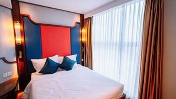 A hotel room with a large bed, a red and blue headboard, and a view of the city through sheer curtains.