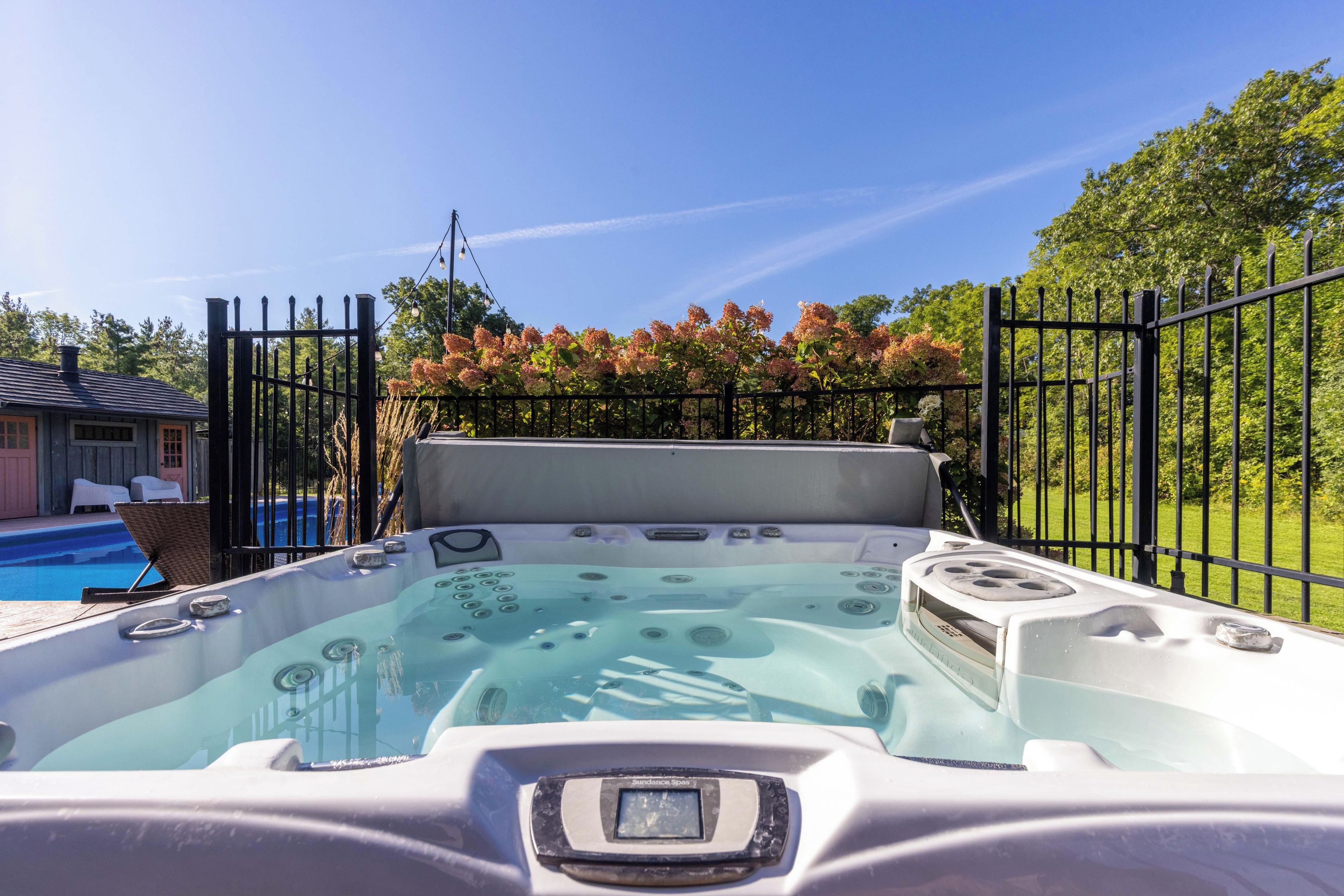 A hot tub with a control panel, surrounded by a fence, with a pool and a building in the background.