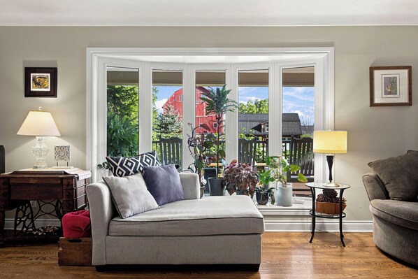A living room with a grey sofa, two side tables with lamps, a wooden floor, and a large window with a view of a red barn and greenery.