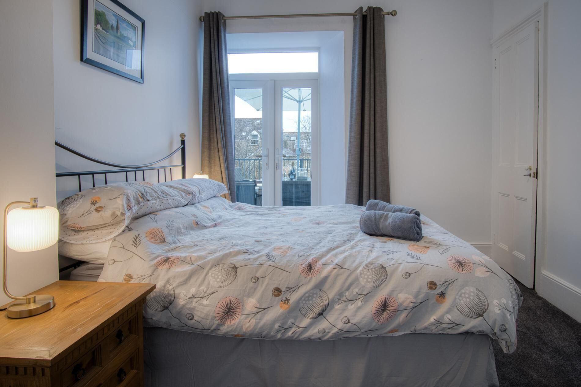 A bedroom with a bed, a bedside table with a lamp, a view through a window, and a door.