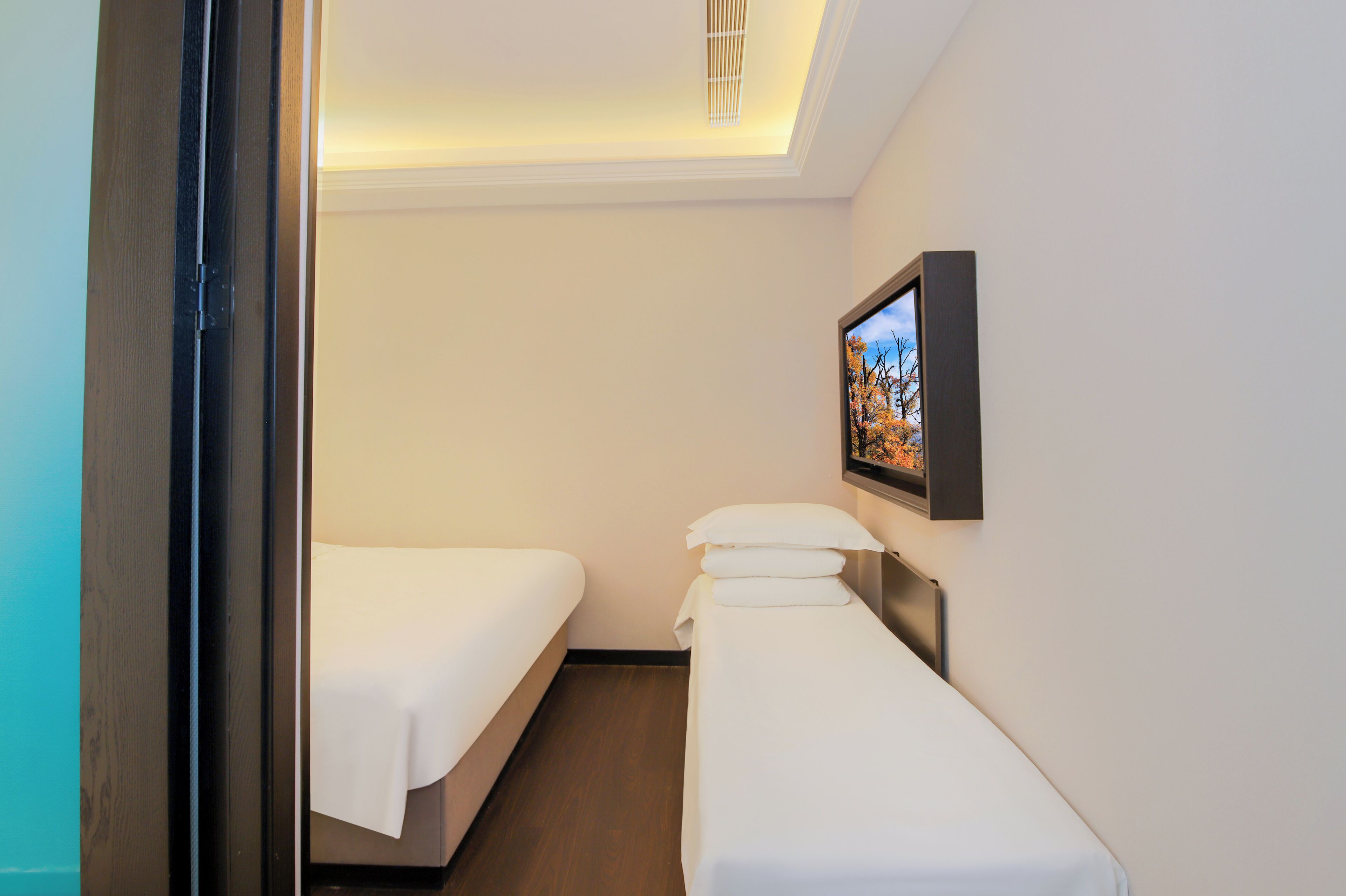 Quarto de hotel com duas camas, uma mesinha de cabeceira, TV fixada na parede e luminária no teto.