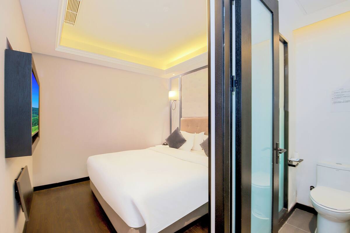 A hotel room with a bed, a bedside table, a lamp, a mirror, and a toilet.