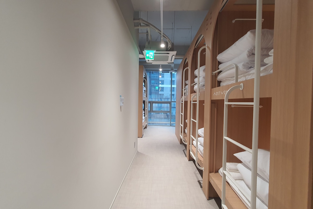 A hotel corridor with bunk beds on the right, a visible exit sign, and a view of the city through the window.