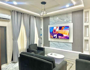 A modern living room with a flat-screen TV, grey curtains, a grey sofa, and a small white coffee table. - 2-bedroom Apartment in serene Opic Estate with AC comfort (Opic)